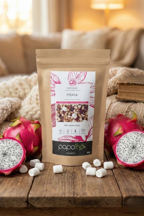 Sachet d'infusion Pitaya Papothe en papier kraft posé sur une table en bois, entouré de fruits du dragon frais roses et de mini-chamallows blancs. Ambiance cocooning chaleureuse avec arrière-plan flou de plaids en maille et lumière douce.